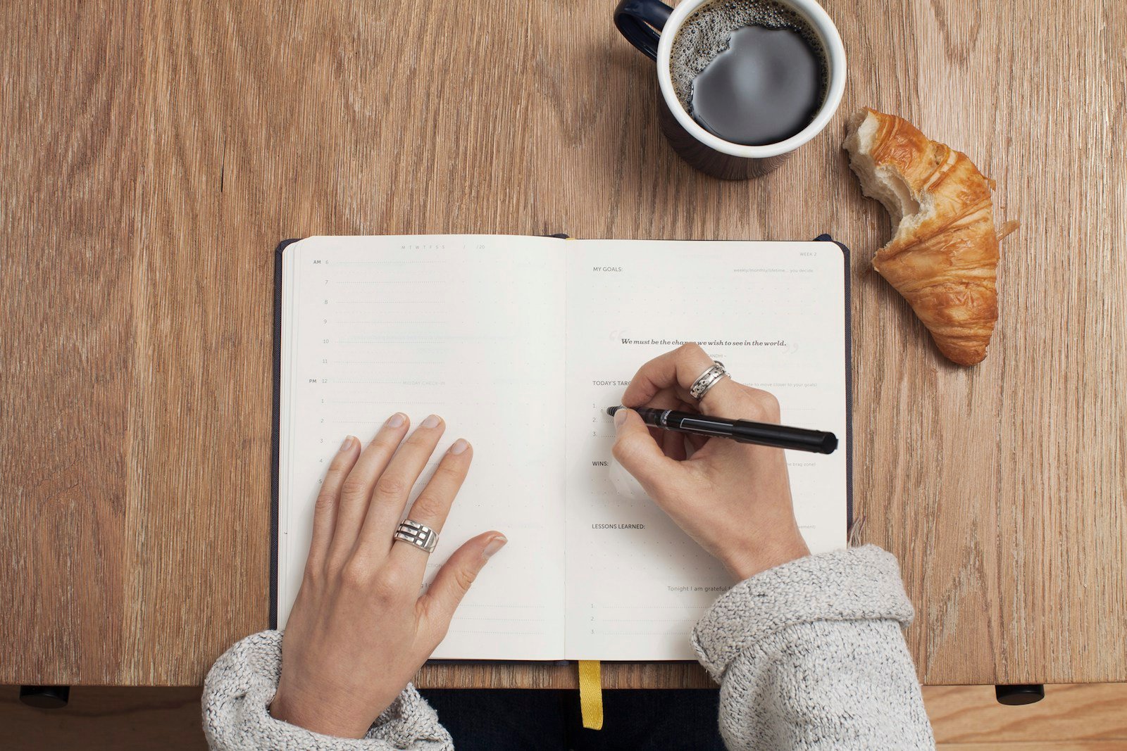 Fotografia ilustrativa de mãos escrevendo em uma agenda aberta sobre uma mesa de madeira.
